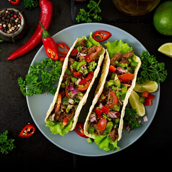 Three Mixteca Grill tacos filled with beef, lettuce, tomatoes, onions, and peppers, garnished with lime and chili.